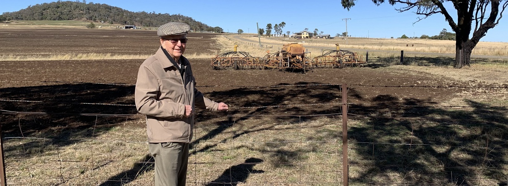 An elderly gentleman stands in front of a paddock where a seeder can be seen in the background An elderly gentleman stands in front of a paddock where a seeder can be seen in the background