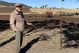 An elderly gentleman stands in front of a paddock, a seeder machine can be seen in the background An elderly gentleman stands in front of a paddock, a seeder machine can be seen in the background